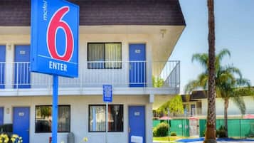Een motel met een opvallend blauw bord waarop nummer 6 staat en een uitnodiging om binnen te gaan. Het gebouw heeft meerdere deuren, genummerd 102, 103, 205 en 206.