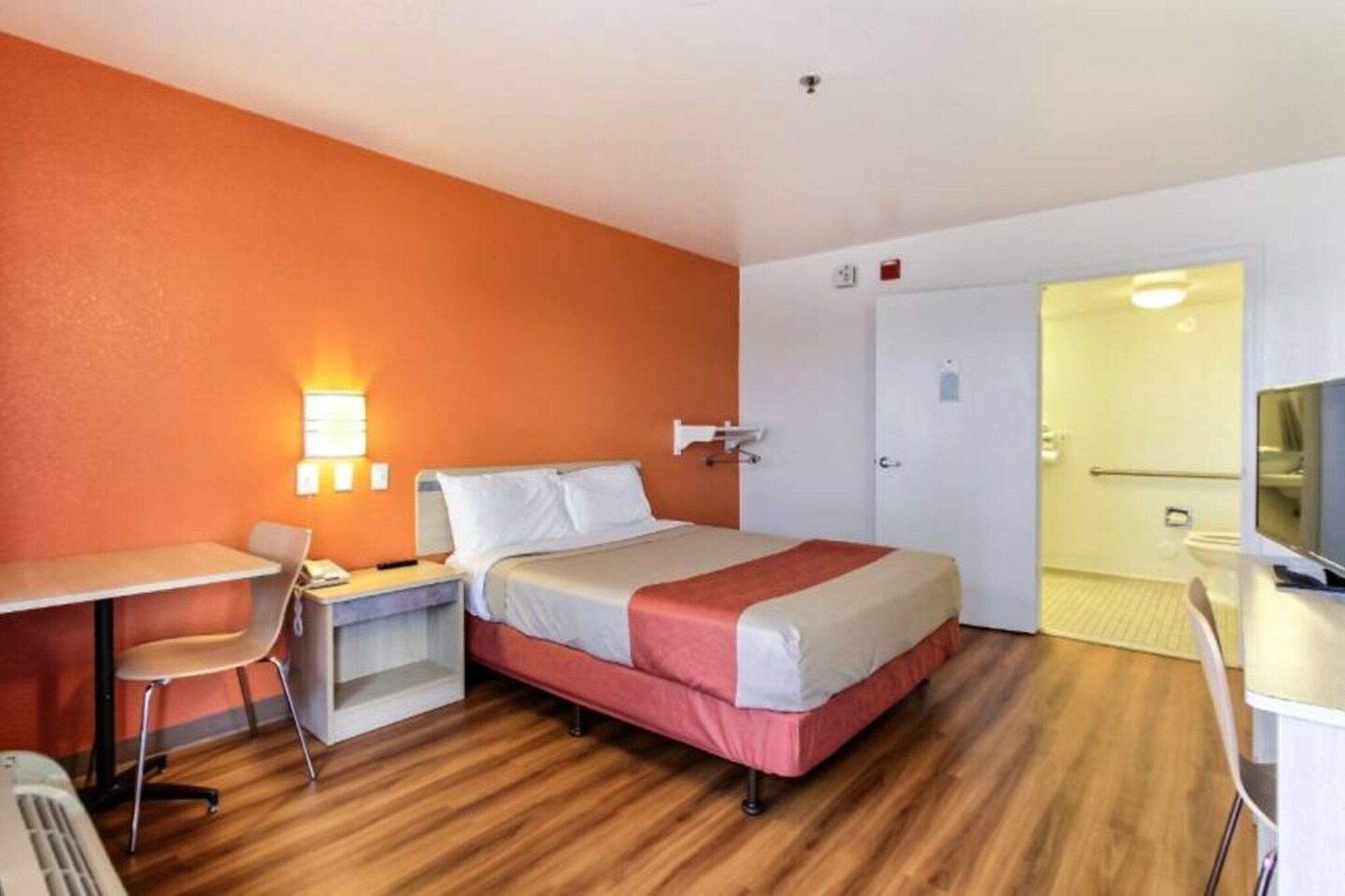 Quarto de hotel com cama, escrivaninha, cadeira, televisão e porta que dá para outro cômodo.