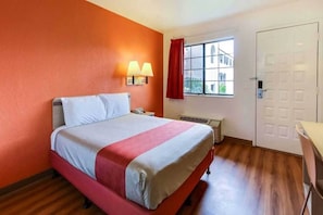 A hotel room with a bed, a window with a view, a door, and a wall-mounted lamp.