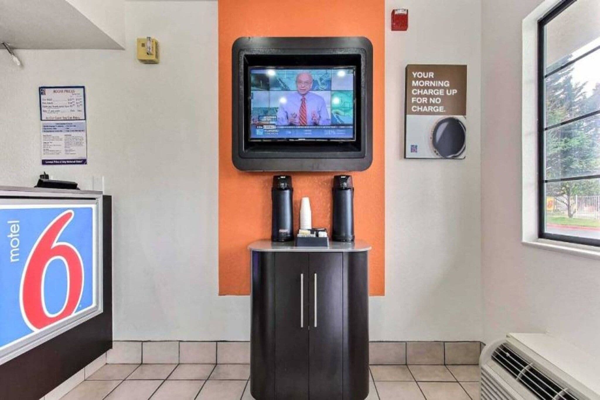 Um saguão de hotel com uma televisão fixada na parede, uma placa indicando cobrança matinal gratuita e uma placa com o número 6 Motel.