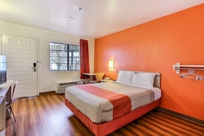 A hotel room with a bed, a desk, a chair, a window with curtains, and a wall-mounted light fixture.