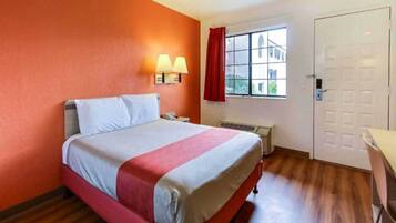 A hotel room with a bed, a window with a view, a door, and a wall-mounted lamp.