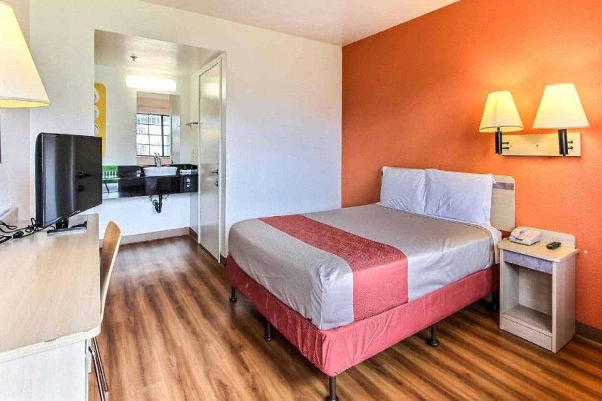 Quarto de hotel com cama, mesa de cabeceira, luminária e escrivaninha com cadeira.