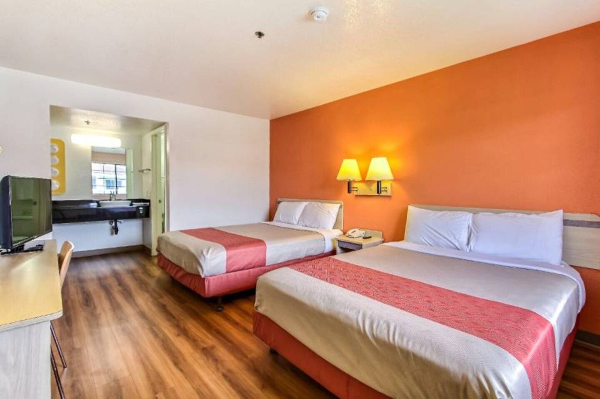 Quarto de hotel com duas camas, uma escrivaninha, uma cadeira, televisão e banheiro visível por uma porta aberta.