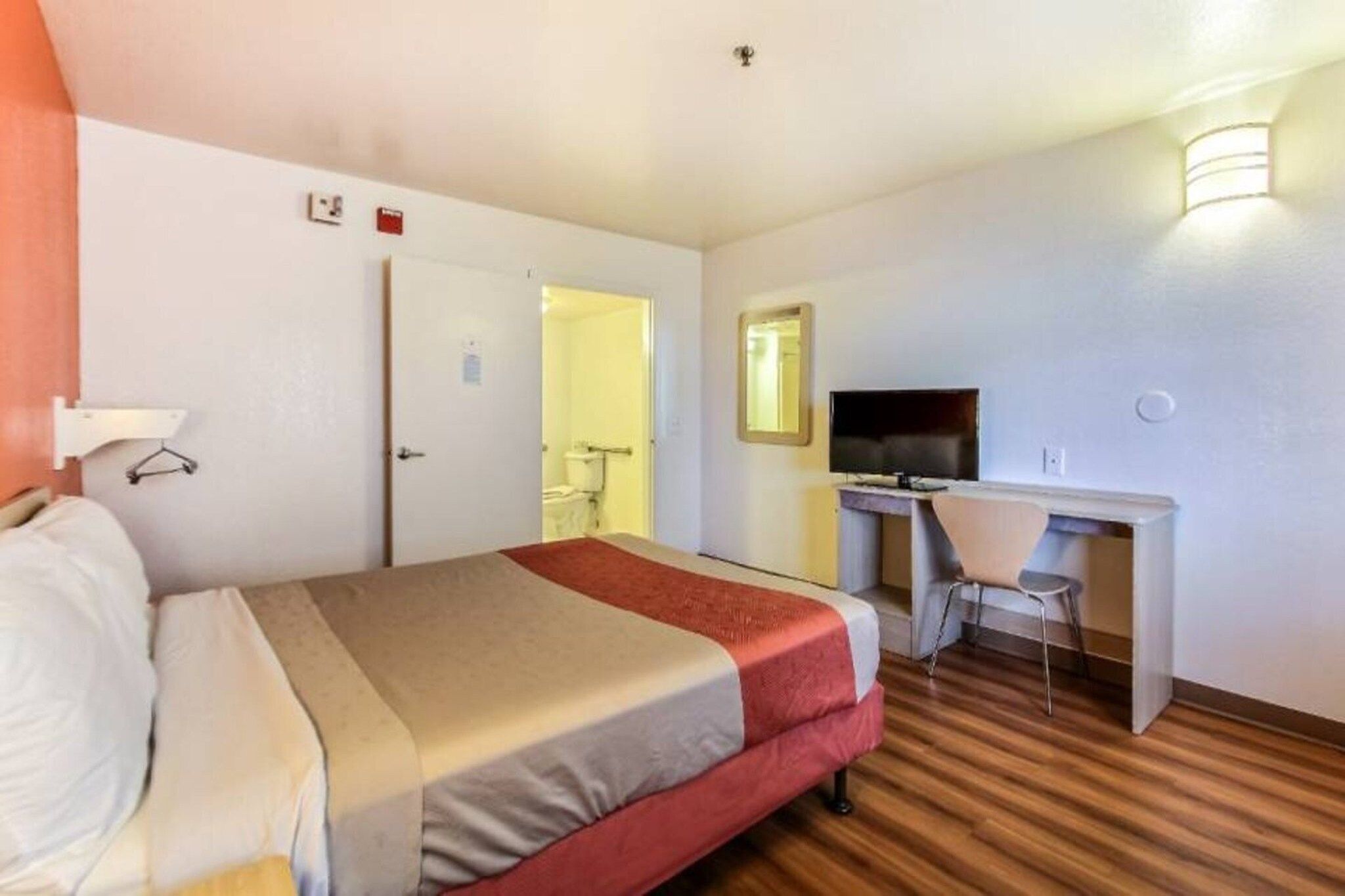 Quarto de hotel com cama, escrivaninha, cadeira, televisão e espelho.