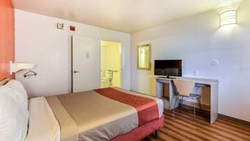 Quarto de hotel com cama, escrivaninha, cadeira, televisão e espelho.