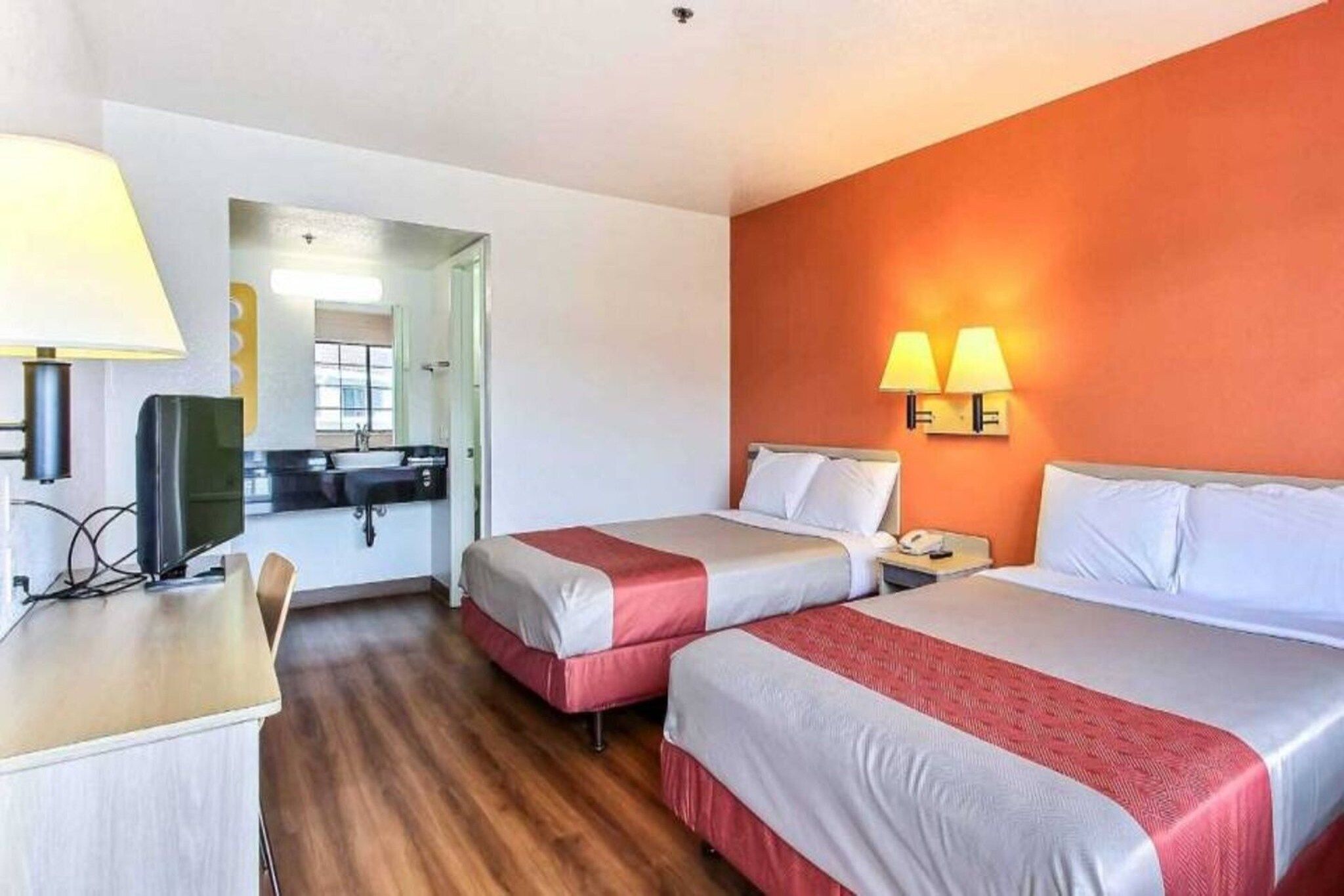 Habitación de hotel con dos camas, un escritorio, una silla, un televisor y un baño visible a través de una puerta abierta.