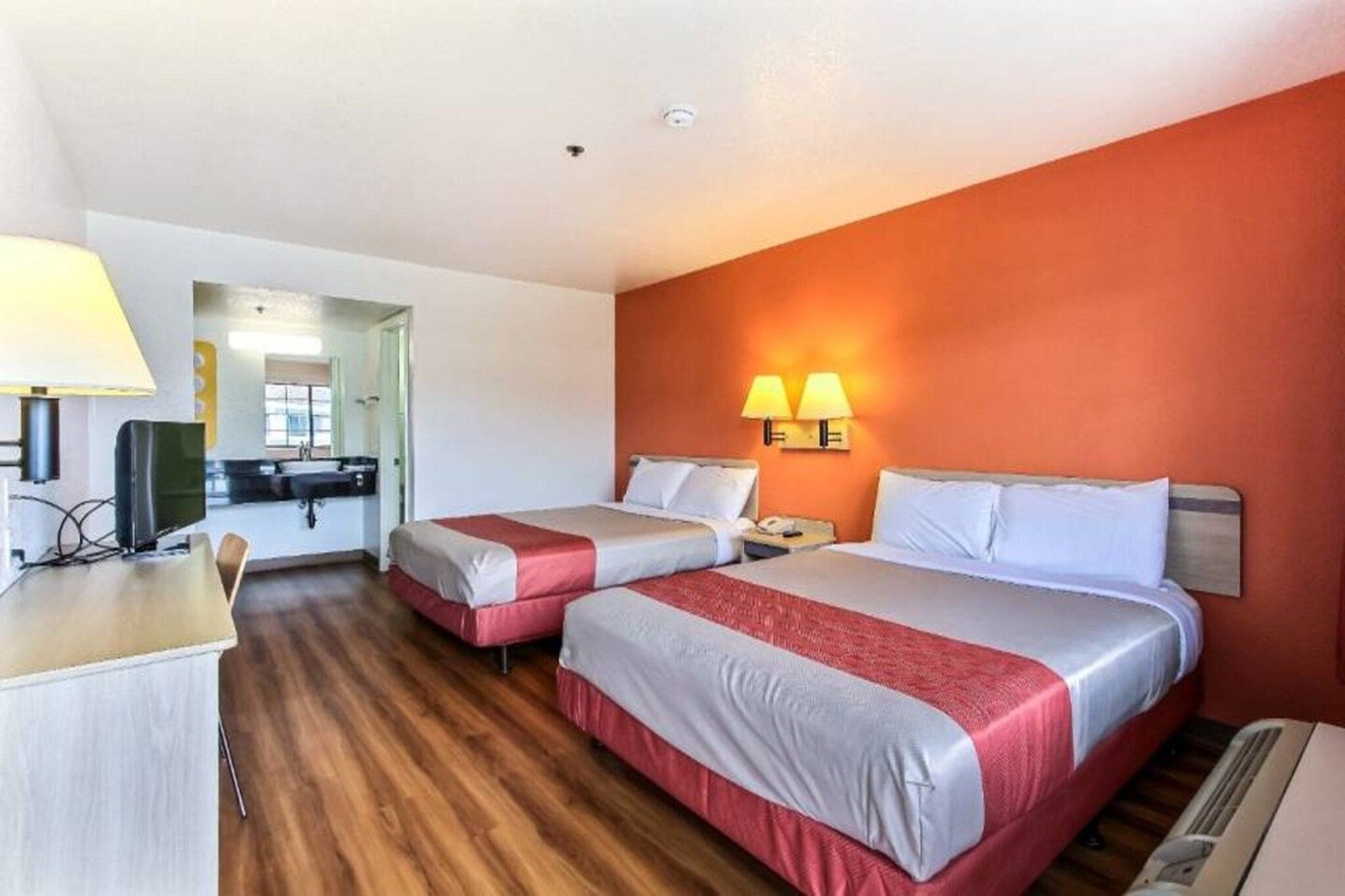 Quarto de hotel com duas camas, uma escrivaninha, uma cadeira, uma televisão e um banheiro visível por uma porta aberta.