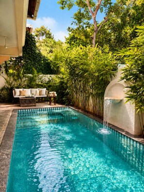Una piscina cristalina con una cascada, rodeada de exuberante vegetación y una zona de estar con mesa y sillas.