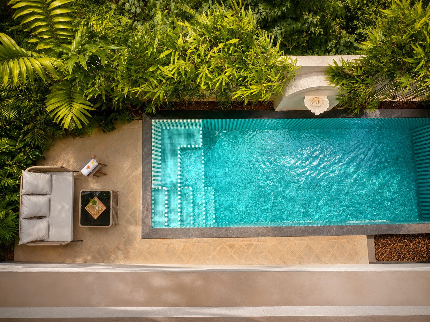 A rooftop pool with a tiled edge, surrounded by lush greenery and a seating area with a towel and a small table.