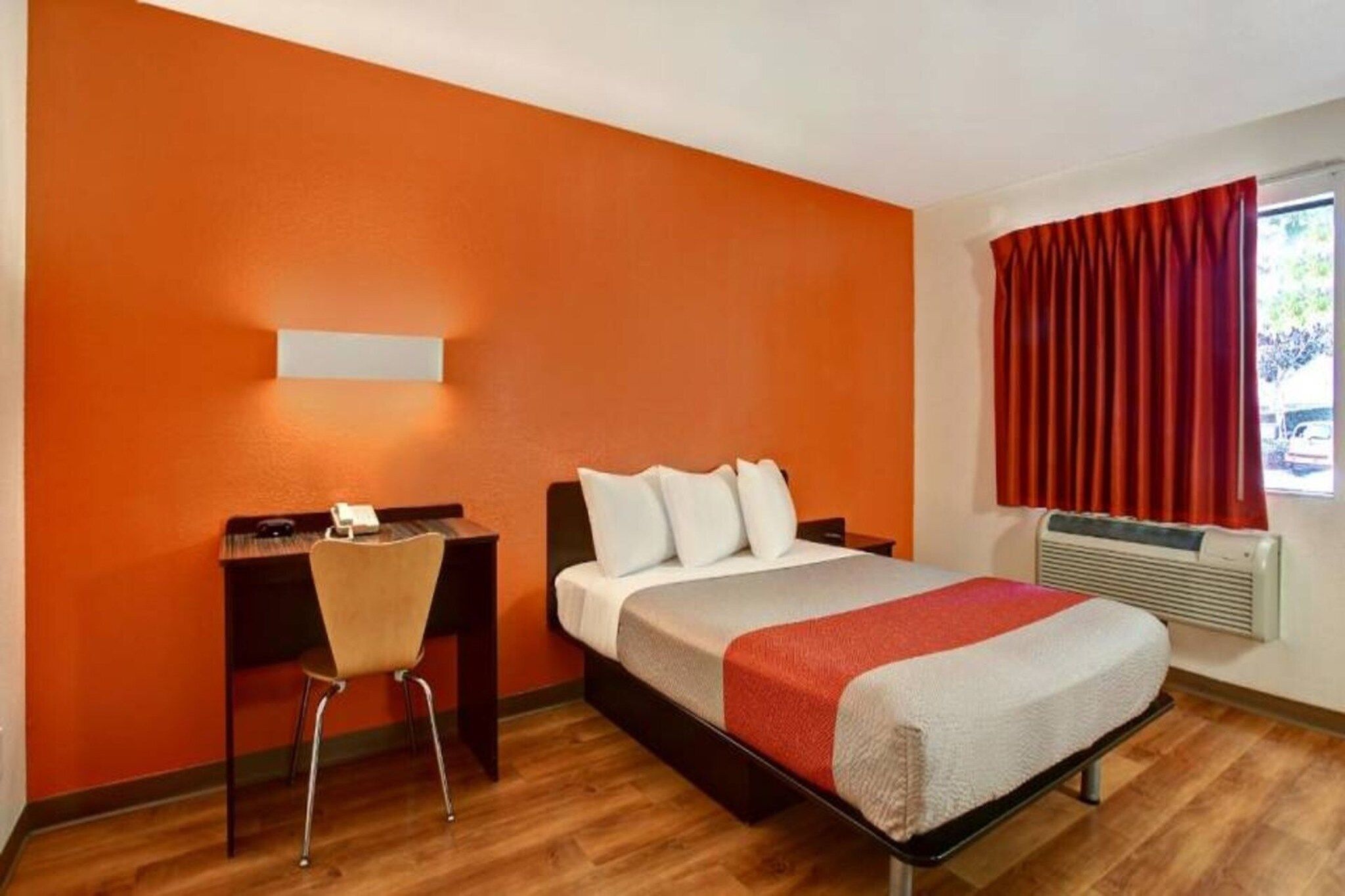 A hotel room with a bed, a desk with a chair, a window with curtains, and an air conditioning unit.