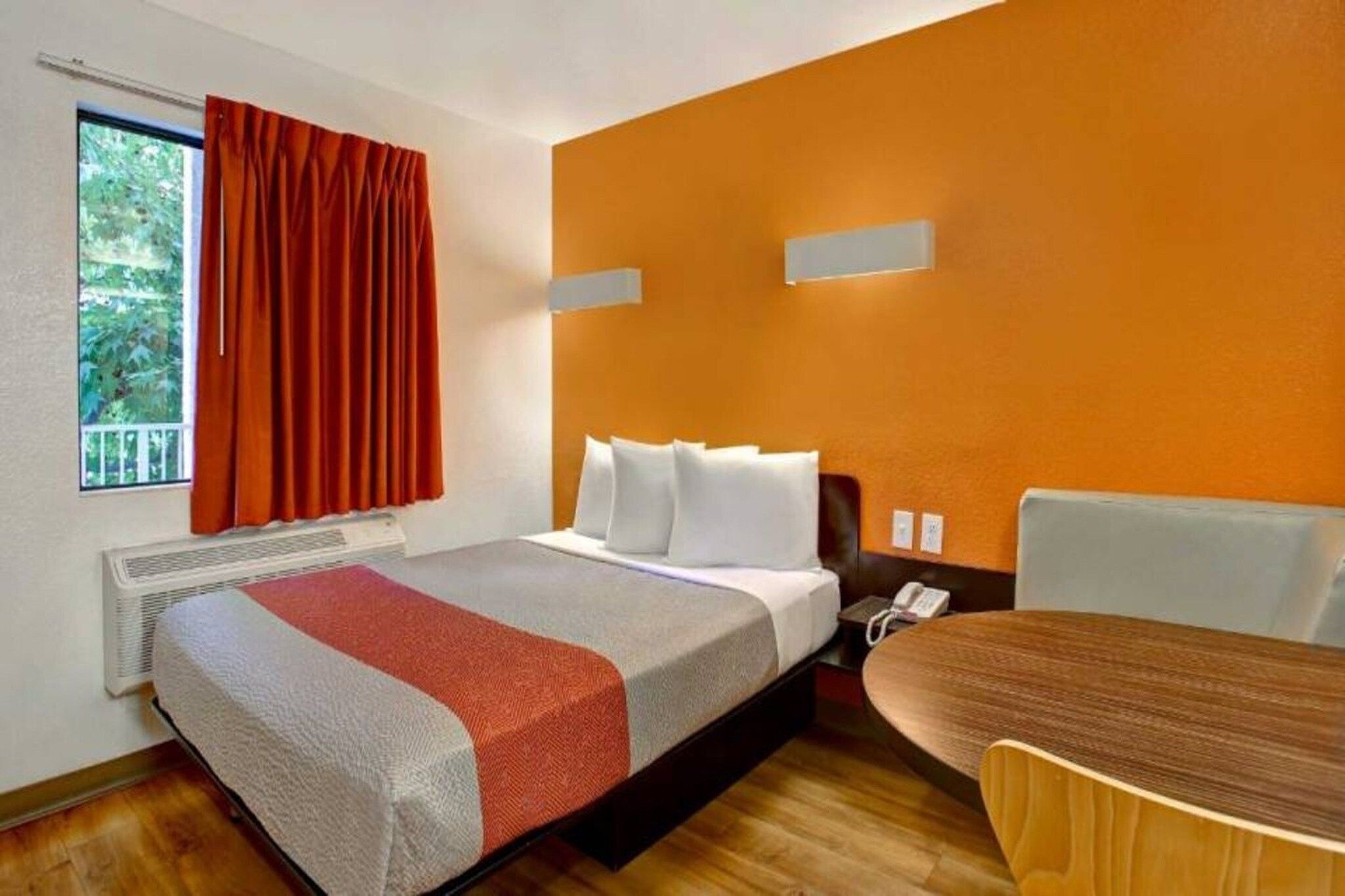 Une chambre d’hôtel avec un lit, une table, une chaise, un téléphone et une fenêtre avec des rideaux.