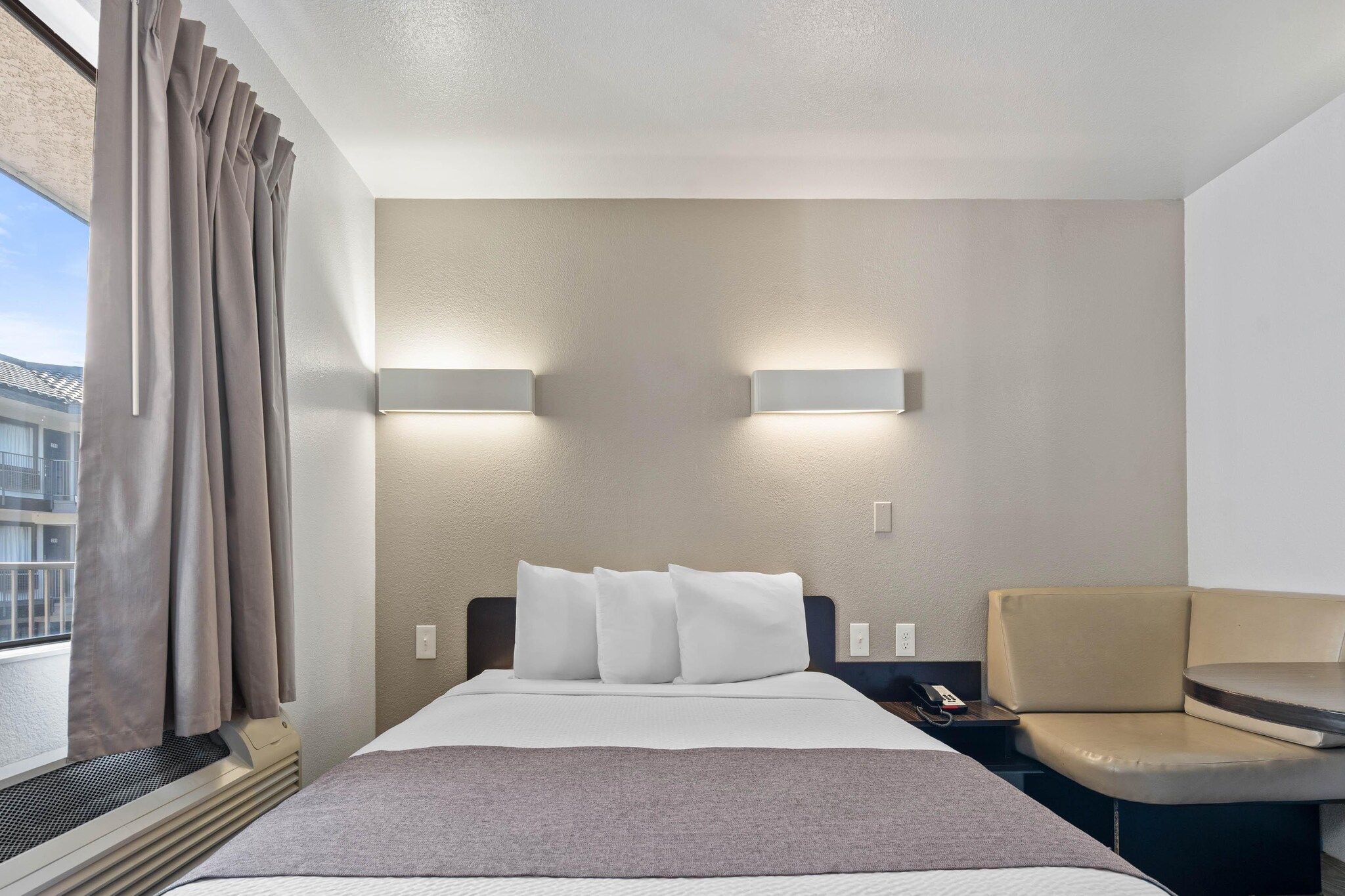 Habitación de hotel con una cama, una silla, una lámpara fijada a la pared y un ventanal con cortinas.