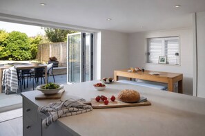 A modern kitchen with a large island, a dining area with a wooden table, and a view of the garden through glass doors. - Wood Cottage (Wadebridge)