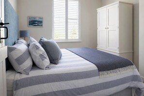 A bedroom with a bed, pillows, a nightstand, a window with blinds, a wardrobe, and a painting on the wall. - Wood Cottage (Wadebridge)