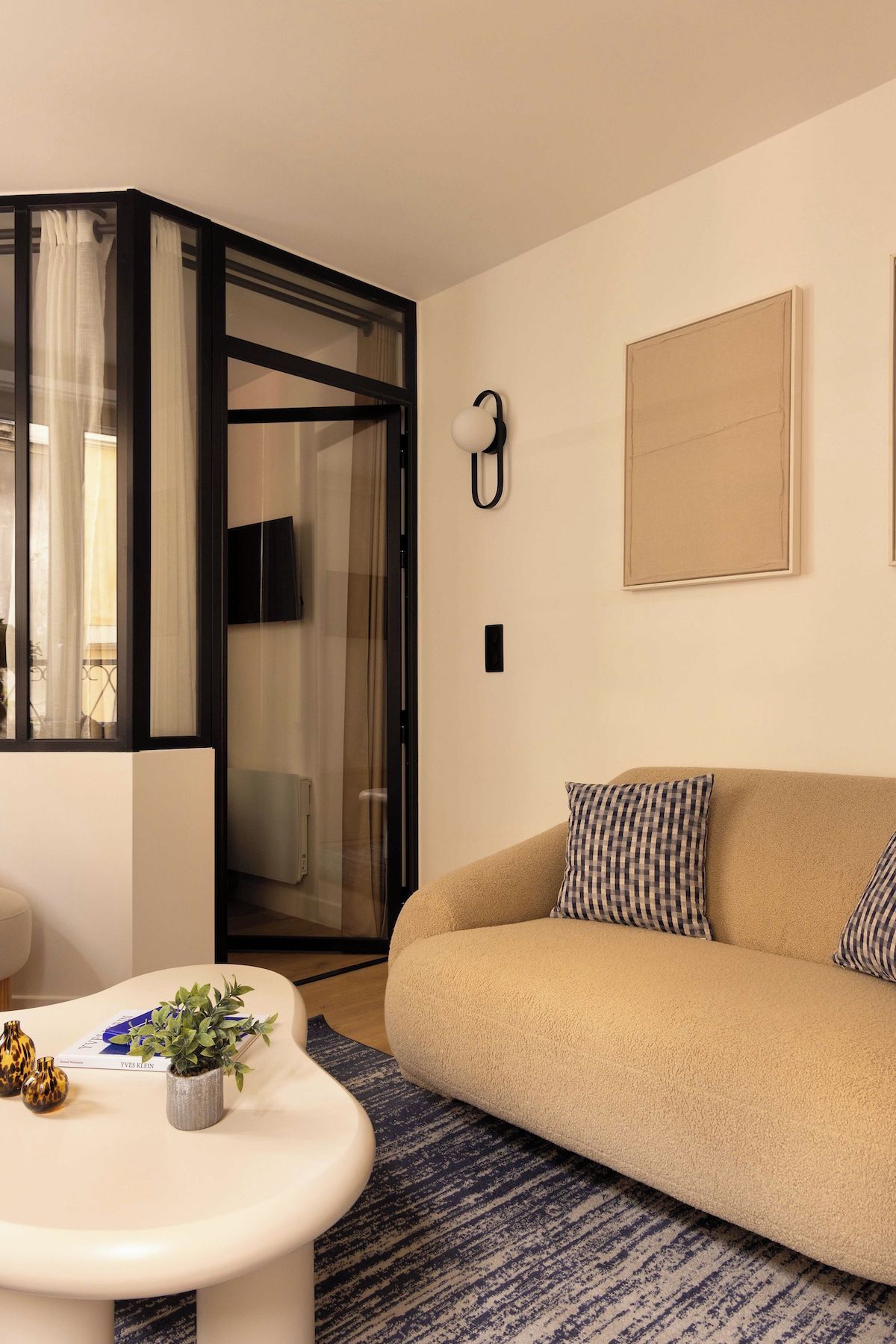 Ett modernt vardagsrum med en beige soffa, ett vitt soffbord med dekorativa föremål, en mönstrad matta och en glasdörr som leder ut till en balkong.