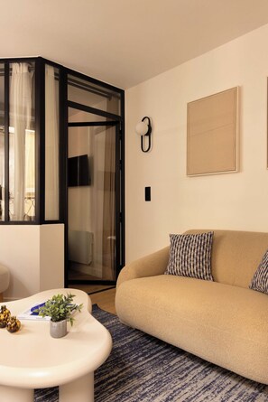 Een moderne woonkamer met een beige bank, een witte salontafel met decoratieve objecten, een vloerkleed met dessin en een glazen deur die leidt naar een balkon.