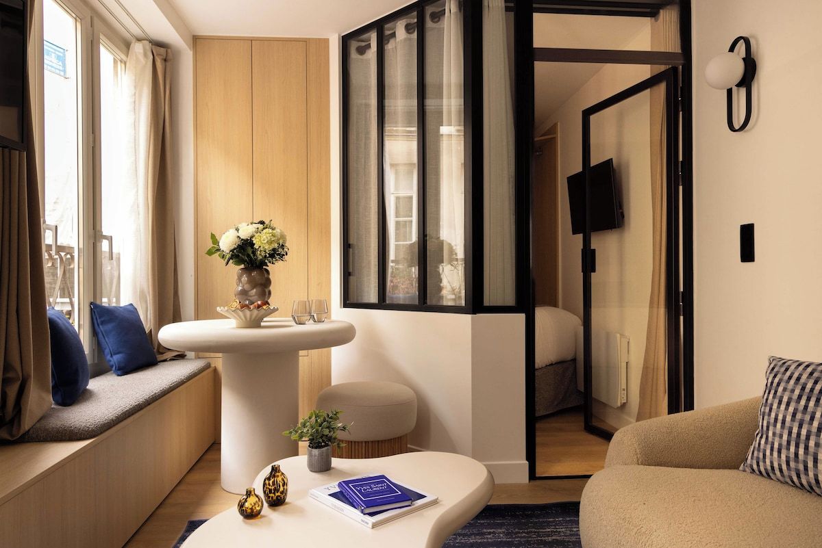 Une chambre d’hôtel moderne avec un canapé, une petite table sur laquelle est posé un livre, un vase avec des fleurs et une vue sur la ville par la fenêtre.