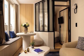 Een moderne hotelkamer met een bank, een kleine tafel met een boek, een vaas met bloemen en uitzicht op de stad door een raam.