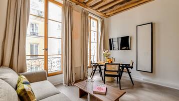A furnished living room with a sofa, a coffee table, a dining table with chairs, a TV mounted on the wall, and large windows with curtains.