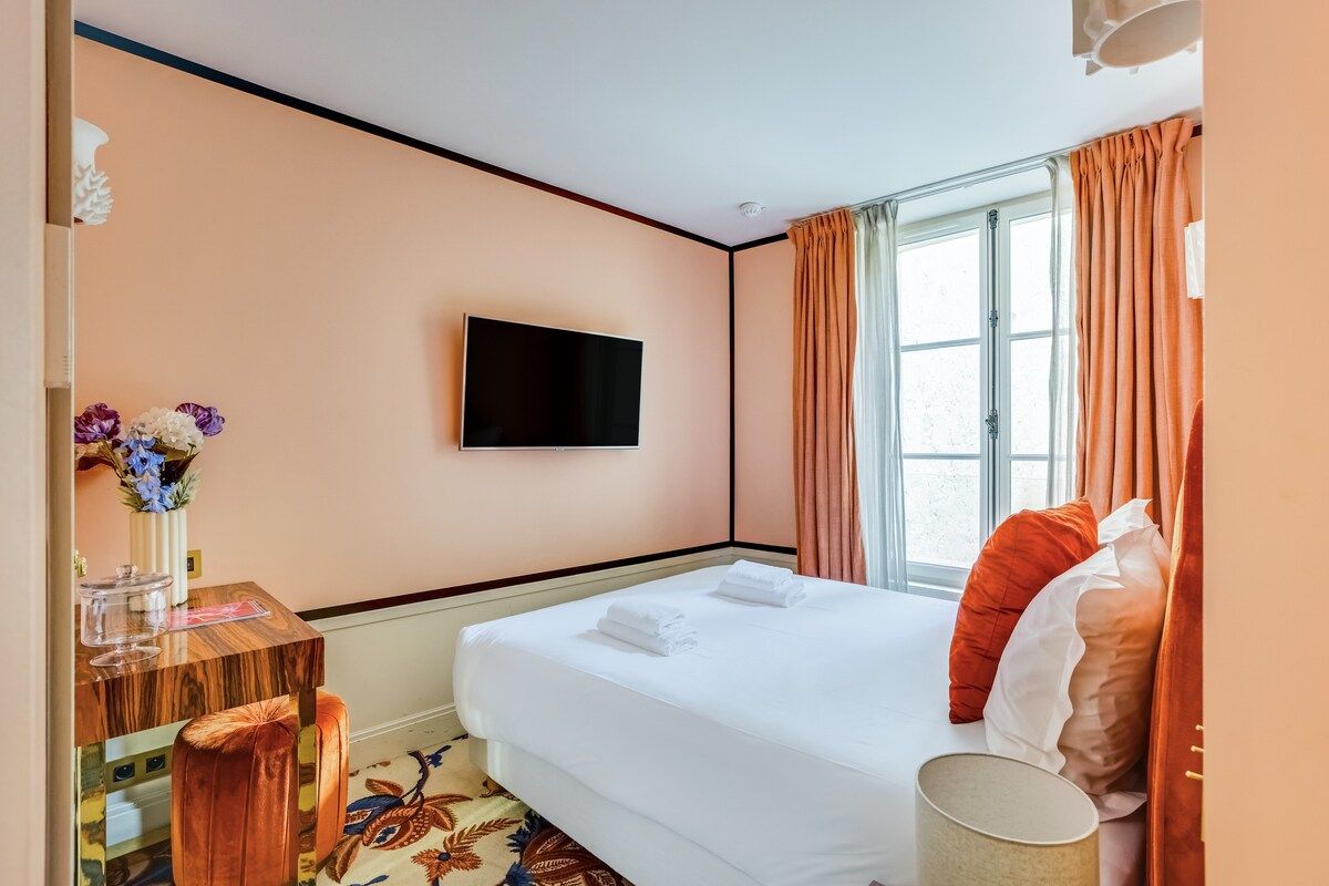 Ein Hotelzimmer mit einem Bett, einem an der Wand befestigten Fernseher, einem Schreibtisch mit Blumen und einem Vorhangfenster.
