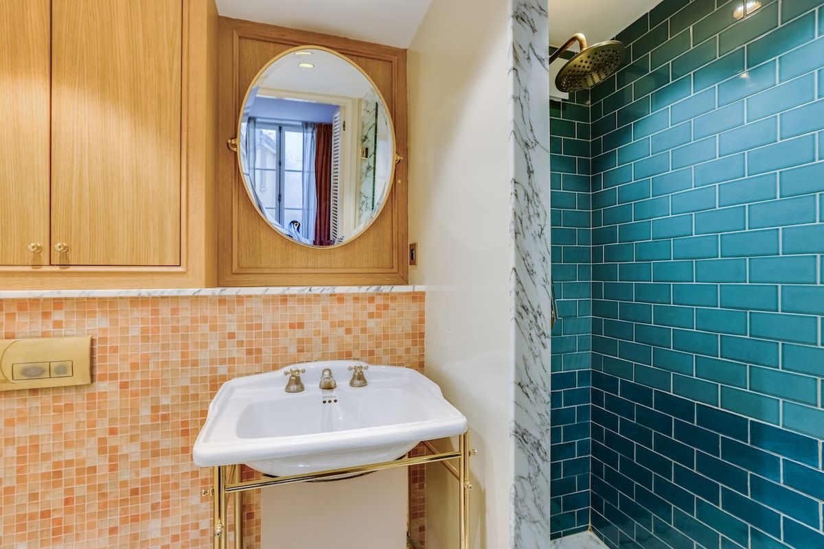 Ein Badezimmer mit weißem Waschbecken, Holdekor-Schränken, einem Spiegel und einer Duschkabine mit blauen Fliesen.