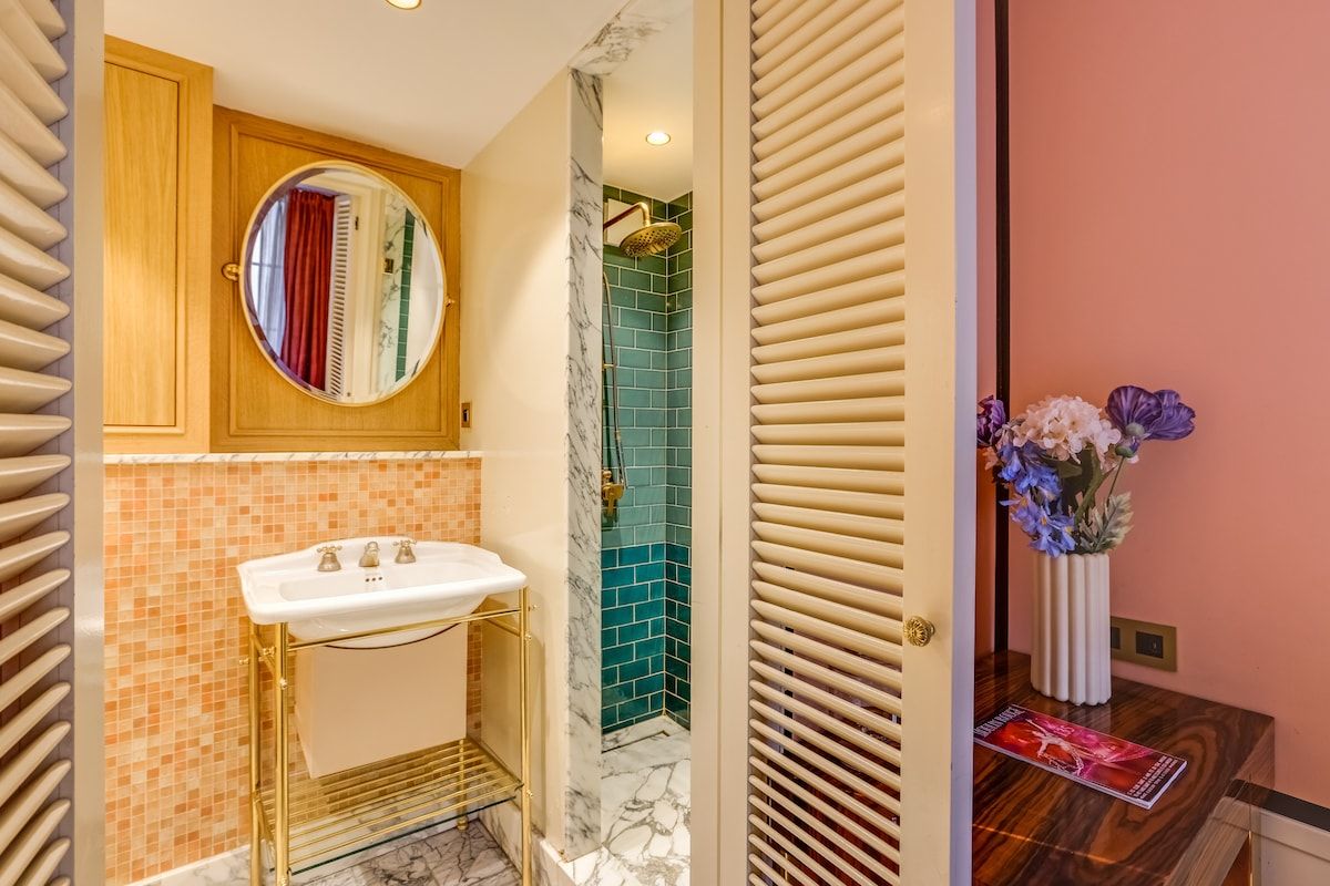 Ein Badezimmer mit einem weißen Waschbecken, einem runden Spiegel, einer Dusche mit goldener Duschbrause und einer Vase mit Blumen auf der Ablagefläche.