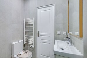 A bathroom with a toilet, a white door, a towel rack, a sink, and a mirror. - Peaceful Studio for a Calm & Soothing Retreat (Paris)
