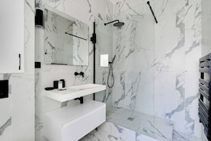 Un baño moderno con pared de mármol, tocador blanco, espejo grande y zona de ducha.