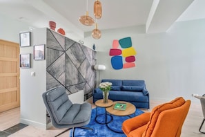 A modern living room with a blue sofa, a grey armchair, and an orange chair. There is a wooden coffee table with a vase of flowers. - Spacious & Modern Apartment - Central Le Marais (Paris)