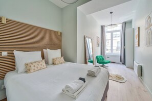 A hotel room with a large bed, a green chair, a mirror, and a view of the outside. - Spacious & Modern Apartment - Central Le Marais (Paris)