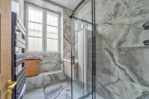 A bathroom with a glass shower enclosure, marble walls, and a window. - Spacious & Modern Apartment - Central Le Marais (Paris)