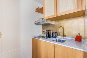 Een keuken met een marmeren aanrecht, een roestvrijstalen spoelbak, houten kastjes, een koffiezetapparaat en een rode zeepdispenser.