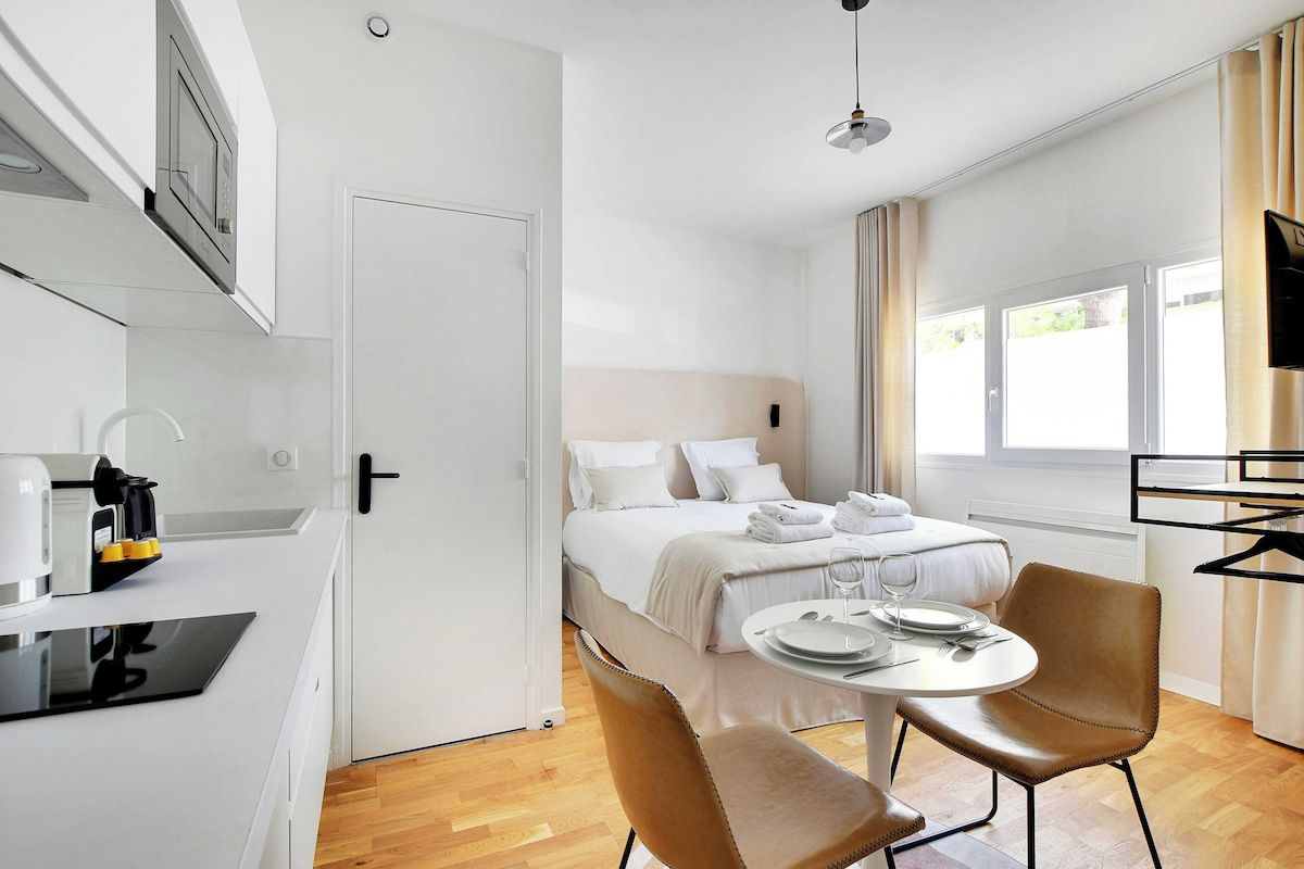 A modern hotel room with a bed, a dining table with chairs, a kitchen area with a coffee machine, and a TV.
