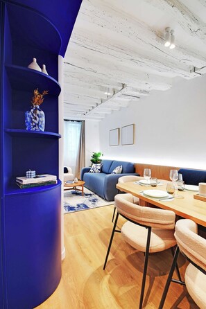 A modern living room with a blue curved shelving unit, a wooden dining table with chairs, a blue sofa, and a white ceiling with exposed beams. - Stunning Flat in the Absolute Centre of Paris (Paris)