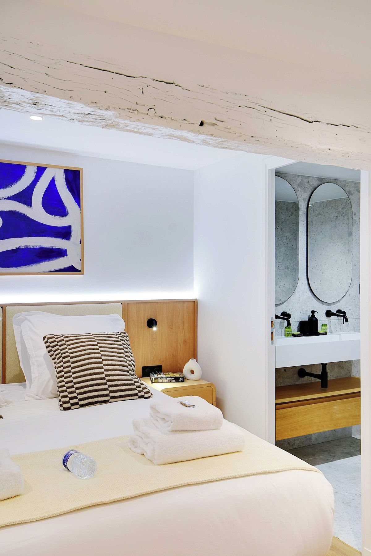 A modern hotel room with a bed, a nightstand, a bathroom with a mirror, and a blue abstract painting on the wall.