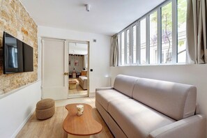 A modern living room with a stone accent wall, a flat-screen TV, a beige sofa, a wooden coffee table, and a large window with curtains. - Parisian Elegance: Modern Flat in Central Paris (Paris)