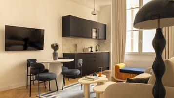Un salon moderne comprenant un coin repas, une cuisine avec des meubles de couleur sombre, un téléviseur à écran plat fixé au mur et un grand lampadaire.