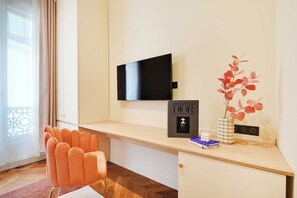A hotel room with a flat-screen TV mounted on the wall, a desk with a book titled 'CHRISTIAN DIOR,' a vase with red leaves, and a chair. - Typical Parisian Flat next to the Louvre (Paris)