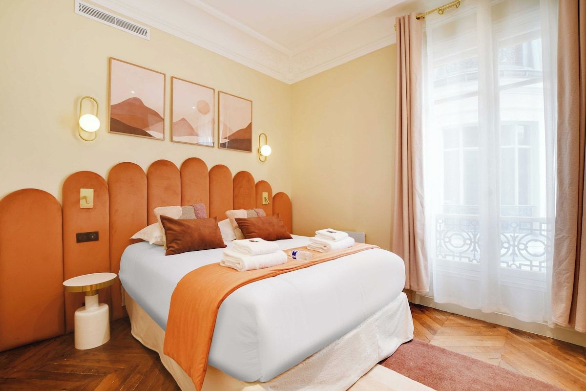 A hotel room with a bed, a bedside table, a wall-mounted light, and a window with curtains.