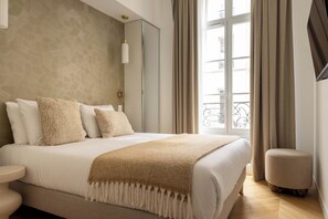 A hotel room with a large bed, a bedside table, a chair, and a view of the outside through a window. - Modern & Refined Flat in Prime Marais Location (Paris)