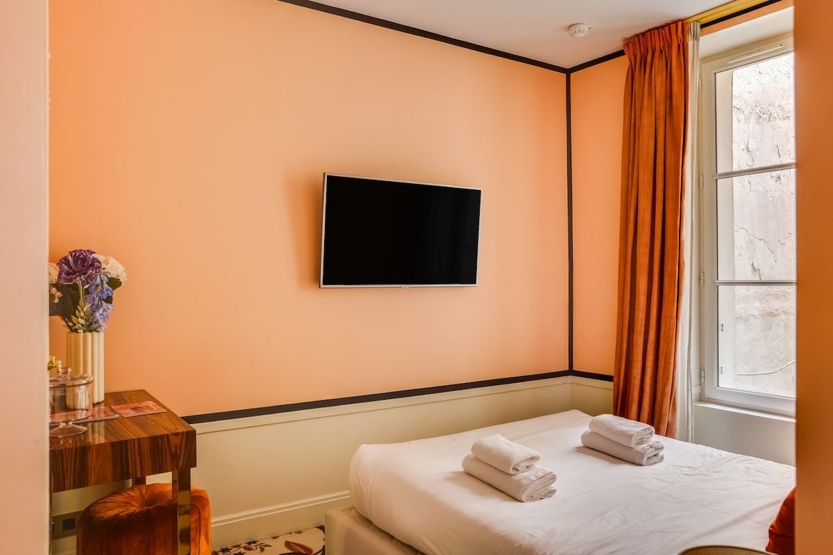 Ein Hotelzimmer mit Bett, Flachbildfernseher an der Wand, Fenster mit Vorhängen und einem kleinen Tisch mit Blumen.