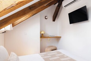 Ein Schlafzimmer mit schräg angesetzter Decke, einem an der Wand befestigten Fernseher, einer Nachttischlampe, einem Regal mit Büchern und einem Bett mit Kissen.