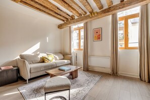 A living room with a sofa, coffee table, and ottoman, wooden beams on the ceiling, and large windows with curtains. - Splendid Duplex overlooking the Heart of Paris (Paris)