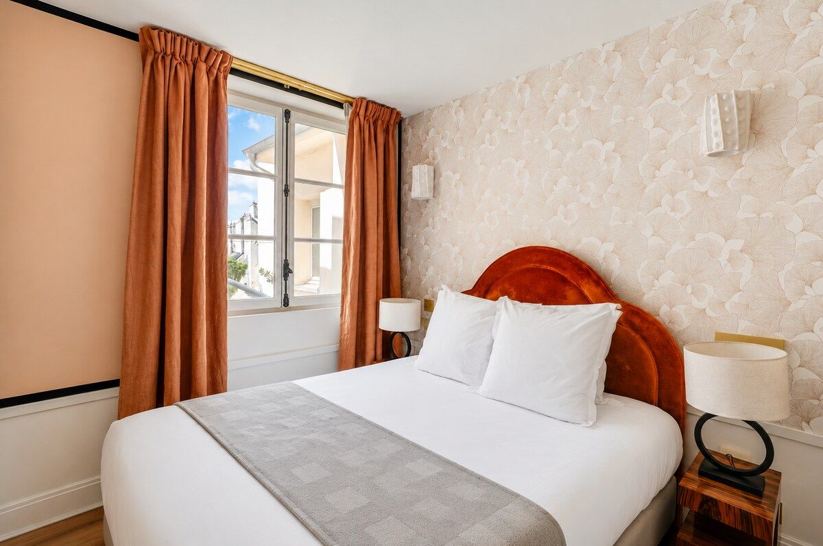 Une chambre d’hôtel avec un grand lit, deux lampes de chevet, une fenêtre avec des rideaux et un papier peint à motifs.