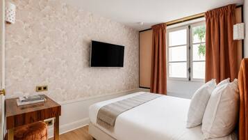 A hotel room with a bed, a flat-screen TV mounted on the wall, a window with curtains, and a small wooden table with a cushion.