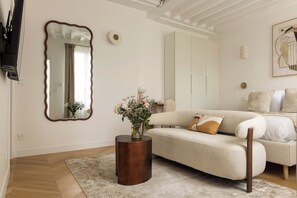 Ein Wohnzimmer mit einer Couch, einem runden Holztisch mit Blumenvase, einem großen Spiegel und einem Bild an der Wand.