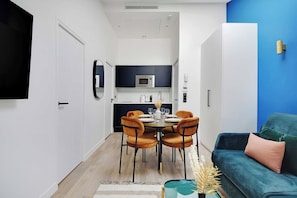 Ein modernes Wohnzimmer mit einem türkisfarbenen Sofa, einem Essbereich mit einem runden Tisch und Stühlen sowie einer Küche mit dunklen Schränken und einer Mikrowelle.