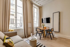 A hotel room with a sofa, a dining table with chairs, a flat-screen TV mounted on the wall, and a window with curtains. - Location, location, location - 1BR near Louvre (Paris)