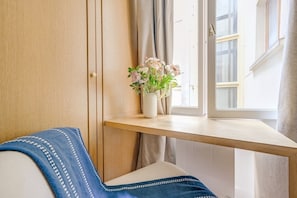 Een kamer met een houten kledingkast, een raam met gordijnen, een klein tafeltje met een bloevas en een blauwe deken op een witte bank.